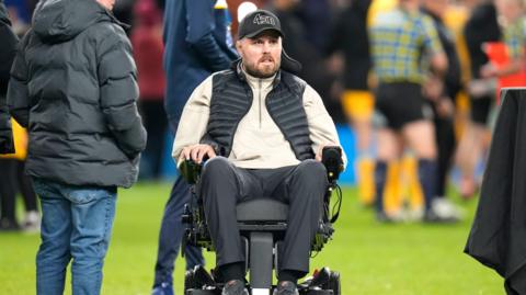 Former Gloucester Rugby player Ed Slater travels across a rugby pitch on a motorised wheelchair. There are people standing around him. He is wearing a black baseball cap, gilet and trousers and a light coloured jumper.