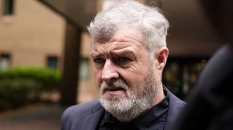 Paul Gallagher leaves Southwark Crown Court in London, where he has denied rape. He is wearing a navy blazer, dark top, has a white short beard, short white hair. 