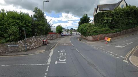 Totnes Road in Paignton at its junctions with Conway Road on the left and Primley Park on the right. A red road closed sign and yellow diversion sign have been set up at the T-junction of Primley Park and are next to a traffic cone. Trees from two gardens are on either side of Totnes Road.