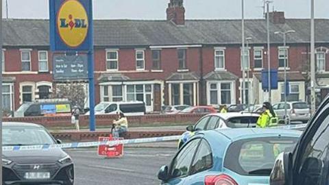 Cars parked on a road with blue and white police tape across the road with ared and white road closed signed. Police officers and a police car is in front of the police tape and a woman with two children sitting on a wall with a blue and yellow Lidl sign in the car park behind her.