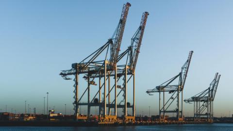 Five enormous container cranes sit on the docks next to the water. The sky behind is completely blue.