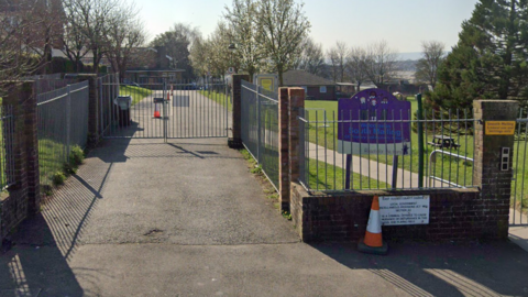 The fence at the front of a primary school. Behind the fence you can see a grass play area and a purple sign, as well as some brick school buildings.