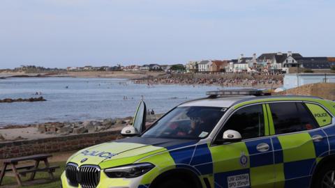 A Guernsey Police car in the usual green and blue police branding parked up on the road over looking a beach with lots of people sat on it and swimming in the sea. There is an officer sat in the car looking out at the people with the car door open. 