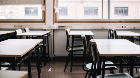 Interior of an empty classroom showing tables and chairs and a window in the background 