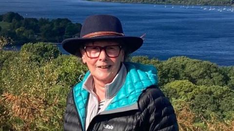 Sally Hall smiles wearing a wide-brimmed hat and a waterproof coat on a hill overlooking a lake. 