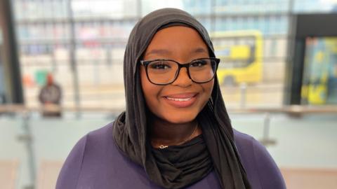 A smiling, young woman wearing black glasses and a hijab. 