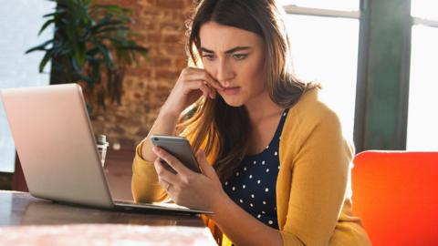 A woman in yellow cardigan looks at a calculator whilst sitting in front of an open laptop. She looks concerned