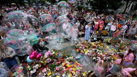 A crowd of people dressed in bright colours blow bubbles into the air over floral tributes. A group of young girls in pink tops stand to one side of the flowers