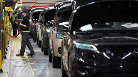 A worker wearing a dark top and a cap is on the left of the photo. Vehicles in a line occupy most of the image.