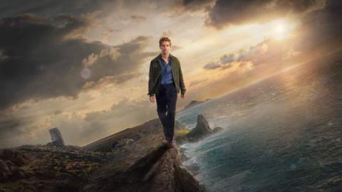 Damien Molony as Det Jim Bergerac walking along a cliff with the sea to his left and Jersey to his right. The sky is cloudy and the sun is setting behind him. He is wearing a green jacket with a blue shirt and black trousers.
