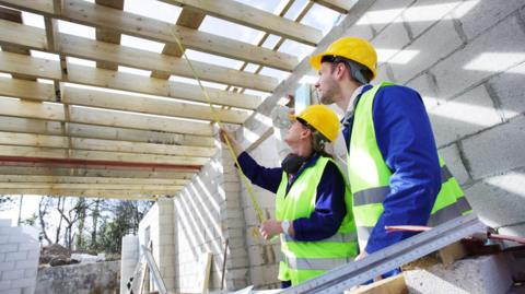 A man and a woman, both wearing dark blue jackets and green high-vis waistcoats, look up towards a partially built wooden roof. The wood in the roof is supported by grey block walls. The woman holds a yellow tape measure with one end poking against a wooden slat in the roof.