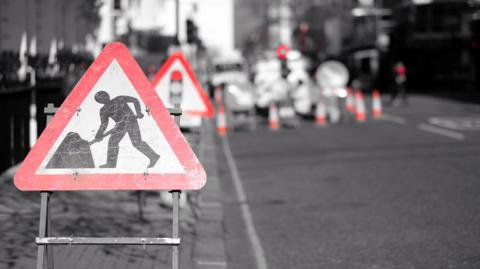 A roadworks sign in the foreground, with a sign for traffic lights further down the road and then a section of road coned off with a set of temporary traffic lights