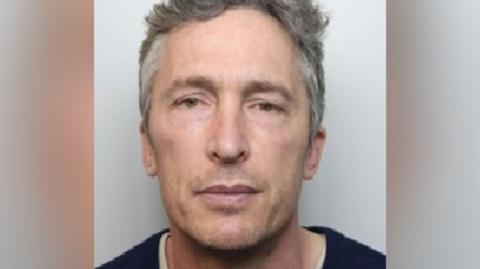 Richard Styles with medium-length grey hair looking at the camera and wearing a black top in a police custody photo