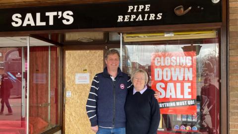 A man wearing a blue gilet and striped shirt stands next to a woman with silver hair, glasses, and a dark jumper with a light collar. He has his arm around her and they both smile at the camera. They stand in front of a shop with red closing down signs in the window. The shop sign, which is black, says Salt's Pipe Repairs.