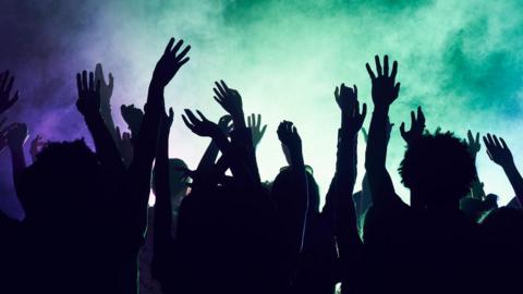 A stock shot of dancers at music event. They are silhouetted with their hands in the air. The room is lit with green and blue light, it is also full of dry ice.