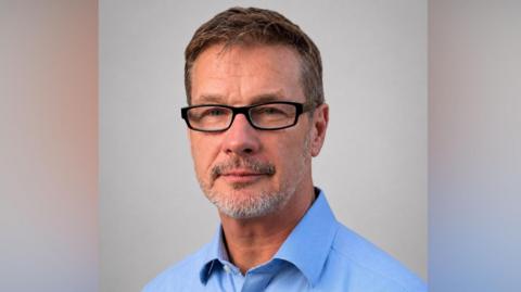 A man in a blue shift with glasses. He has grey stubble. 