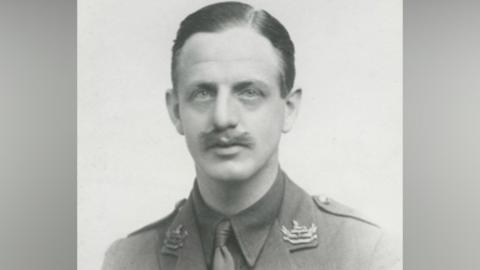 A black-and-white image of a World War One soldier with moustache and brown hair combed to the side. He is wearing an army uniform with shirt, tie and jacket and insignia on the collar.