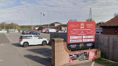 Entrance to Sandal RUFC on Standbridge Lane in Wakefield, with a sign bearing the club's name on the left, and a car park and rugby pitches in the background.