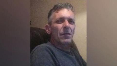 A man in his mid-fifties with grey hair. He is sitting in an armchair and squinting at the camera.