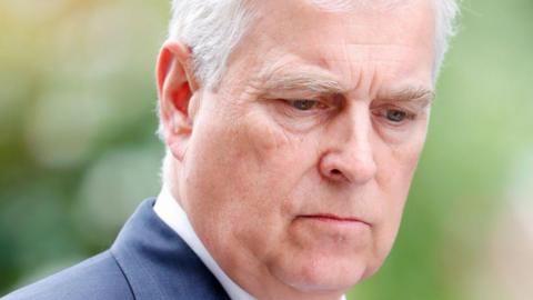 Prince Andrew, who has white hair and wears a blue jacket, looks down.