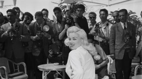 Black and white image of Diana Dors wearing a white blouse, with a large number of photographers behind her.