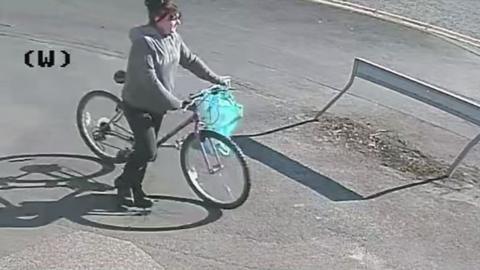 Image of the suspect a woman with dark hair wearing a grey hooded top and dark trousers pushing a bicycle with a blue bag on the handle bars 