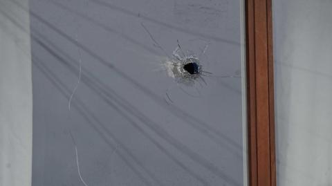 The photo is a close-up of a bullet hole in the window of a house. The window frame is brown wood. 