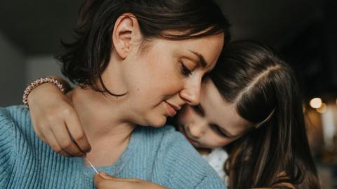 A mother and daughter together. The young girl has her arms around the older woman playing with her necklace.