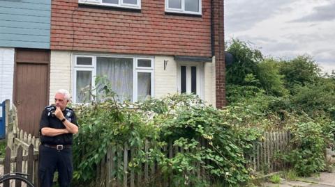 A uniformed police officer stands outside the gate of an end-terrace house. He has one arm crossed across his waist with his other hand to his mouth as if he's thinking. The house is made from bricks, with the bottom half painted cream and the top half tiled. A small front garden is fenced in and the fence is covered with overgrown foliage and shrubbery. 
