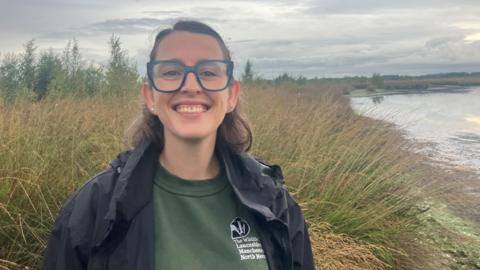 Jenny Bennion is pictured, smiling as she stands on the peatland. She has shoulder-length brown hair and is wearing glasses and a khaki-green sweatshirt with a Lancashire, Manchester and North Mersey logo and a dark-coloured rain coat.