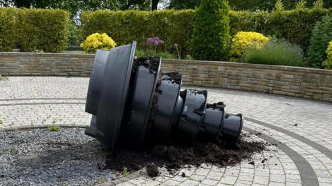 A large black plastic tiered planter has been tipped on to its side in the middle of a circular paved area of a garden. Green hedges and bushes ring the wall at the perimeter.