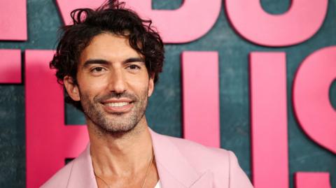 Justin Baldoni in a pink jacket, smiling and posing at the It Ends With Us premiere in 2024 in front of a dark background showing part of the film's title in giant pink letters