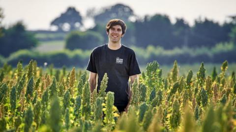 Man with dark hair wearing a black T-shirt.  He is standing in the middle of a field with green crops.  There are trees and bushes in the background.