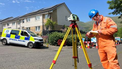 A man in hi vis looks at a notebook, with a measuring device beside him. Police officers are visible in the background