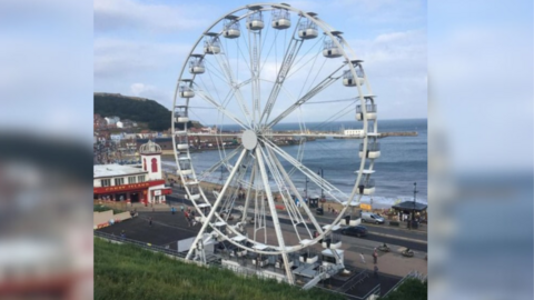 A large Ferris wheel occupies a site along Scarborough sea front. The wheel is white, with evenly spaced gondolas. Behind it, a view of the sea, the beach and the coastline can be seen, and to the left, a white building housing an amusement arcade.