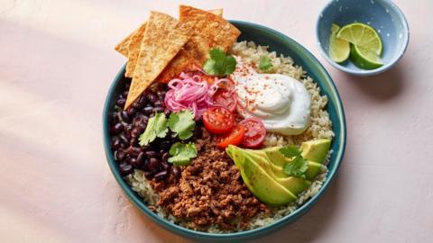 A bowl with beef mince, black beans, sliced avocado, tomatoes, sour cream and tortilla chips on a bed of white rice