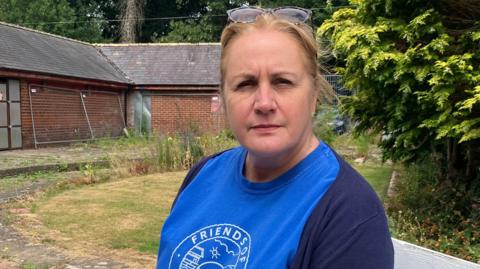 Leonie stands for a photo. She is stood in the grounds of the former Otley Lido. She wears a blue t-shirt which reads 'friends of Otley Lido', under a navy cardigan. She has blonde hair tied up.