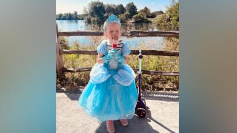 A girl in a blue dress with purple shoes holds an ice cream standing next to a scooter.