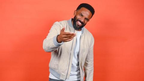 Craig David smiles and gestures to the camera - he has short black hair and a beard, and wears a cream jacket