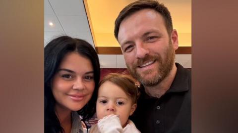 Jody is holding up her daughter and standing next to her partner in this selfie photo. 