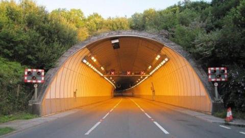 Entrance to the Saltash Tunnel. The entrance is lined with trees. Lights are on inside the tunnel. The gantry above the carriageways have red Xs on them.