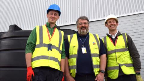 Workers at Rheinmetall BAE Systems in Telford next to the smart water butt. The three men, wearing hi-vis vests and hard hats are pictured smiling in front of a white brick building. 