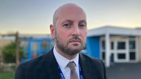 Headteacher of Queensway Primary School, Mark Duce looking at the camera. He wears a suit and tie. The school, which is a blue building, is in the background.