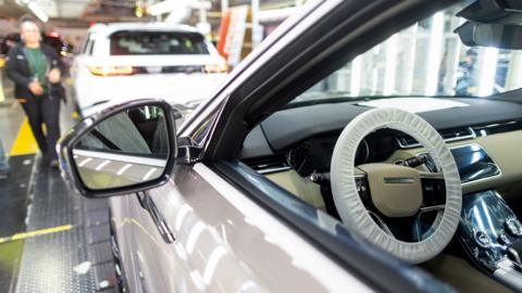 An employee is seen walking in the background behind a close up of a car wing mirror and interior showing the steering wheel and white bodywork, on the production line at JLR's Solihull factory in 2023.