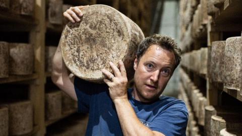 A man stands in a warehouse with shelves stacks with large wheels of a cheddar cheese, on of which he is holding on his shoulder.