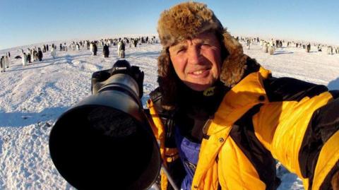 Doug Allan - selfie of man in puffer jacket and woolly hat, holding large camera in a snowy location surrounded by penguins