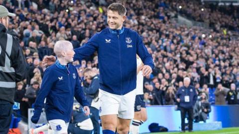 Mason, who is wearing an Everton FC kit, is walking on to the pitch alongside James Tarkowski. Crowds of spectators can be seen in the background. 