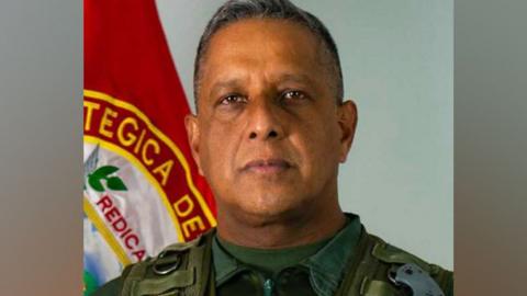 Javier Marcano Tábata stands in front of a flag. He is uniform and looks at the camera.