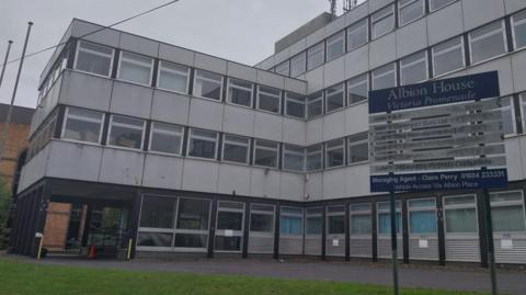 A large grey office block. The main part is four-storey, with a smaller three-storey section attached. There is a blue and silver sign out the front that says Albion House.