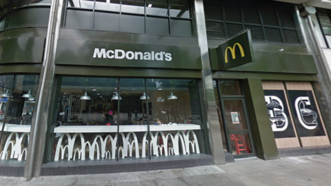An external view of the McDonald's branch, showing white benches and seating inside the restaurant. The outside of the restaurant is dark green.